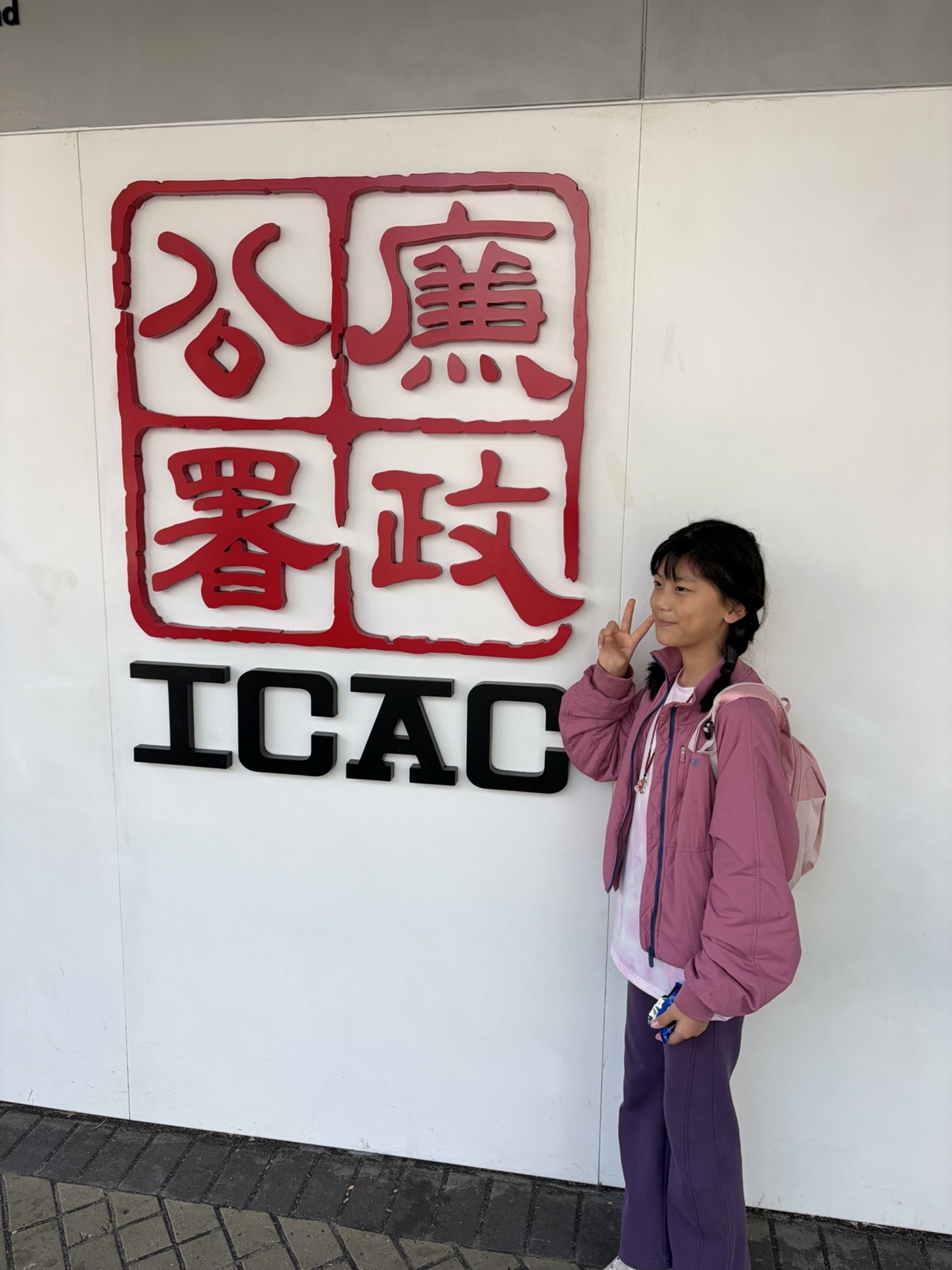 女孩背着粉色背包，在廉政公署 ICAC 标志前留影